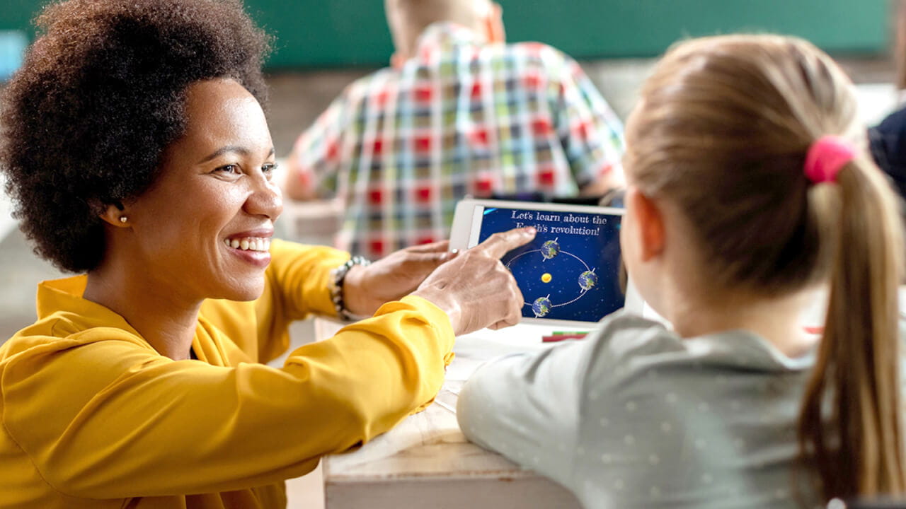 A teacher in a yellow blouse, using Lumio on a tablet, engaging a student with an interactive lesson about the Earth's revolution in a classroom setting.