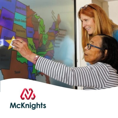 A resident at a senior care facility, along with her aid, using a star shape stamp on an interactive whiteboard, with an image of a map of the United States on it. The McKnights Senior Care logo appears at the bottom of the image.
