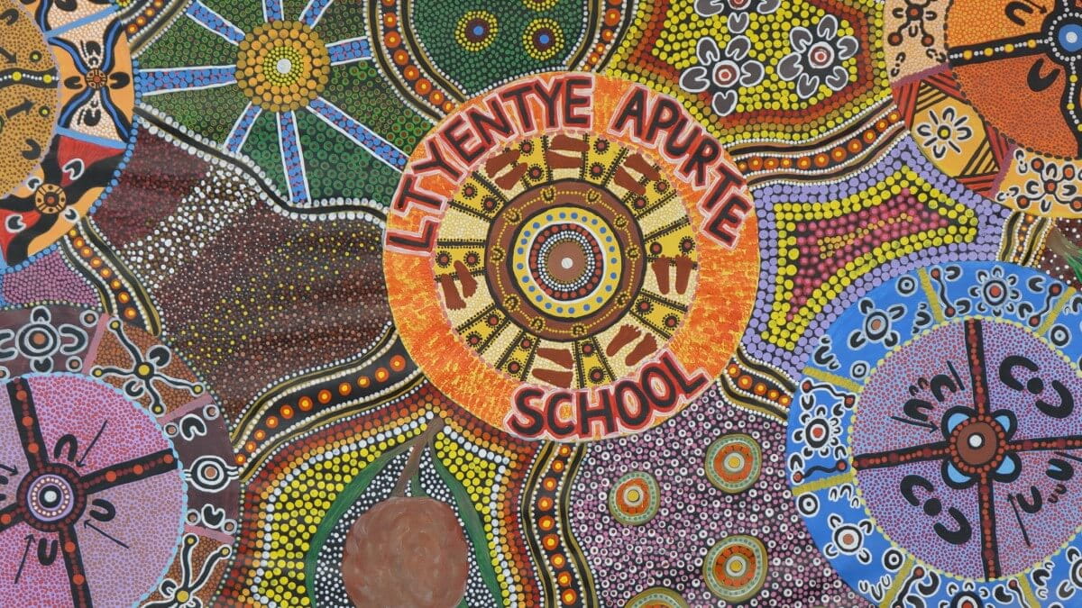 Vibrant Aboriginal-inspired artwork featuring intricate dot patterns, symbols, and a central motif that reads 'Ltyentye Apurte School' surrounded by radiant designs.