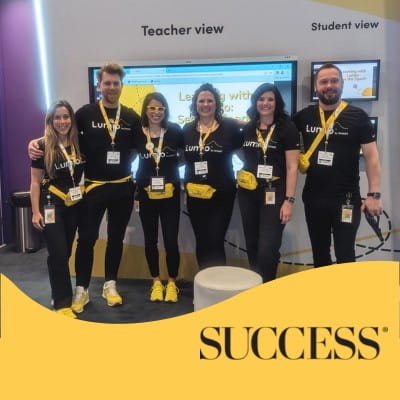 Six members of the Lumio team standing together at the Lumio booth at an education trade show. The yellow banner at the bottom reads “Success.”