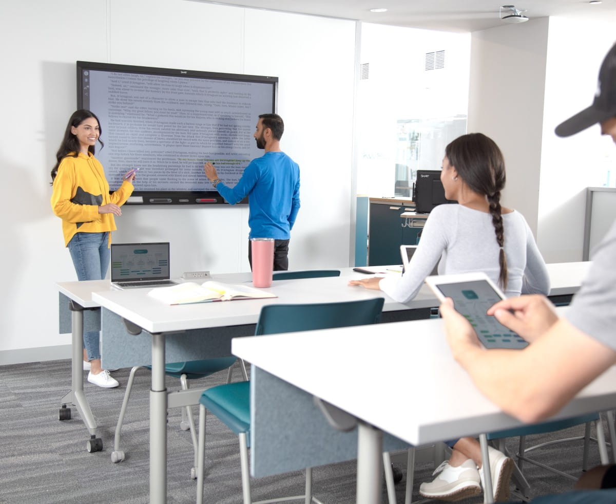 A vibrant classroom setting with ample lighting shows a student actively interacting with a SMART board, which is showing a text document. The teacher, with a cheerful expression, stands close by, while other students, seated at their desks.