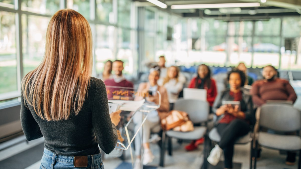 A female professional standing at a podium and presenting to a room of people.