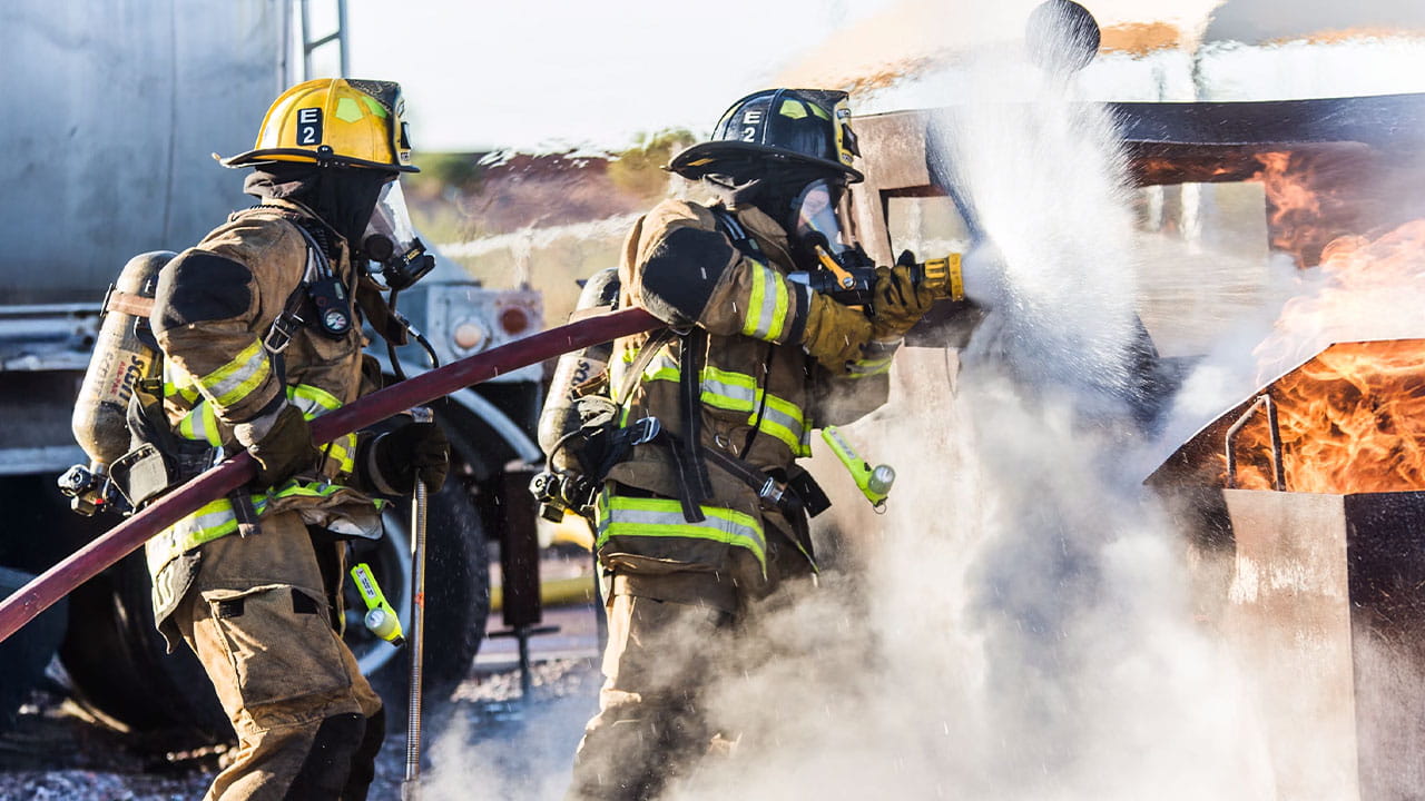 Two firefighters in full protective gear using a fire hose to extinguish flames during a live fire training exercise.