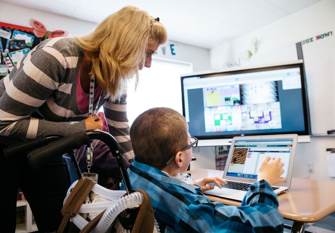 A teacher supports a student in a wheelchair using a laptop in front of an interactive SMART Board, highlighting inclusive education technology.