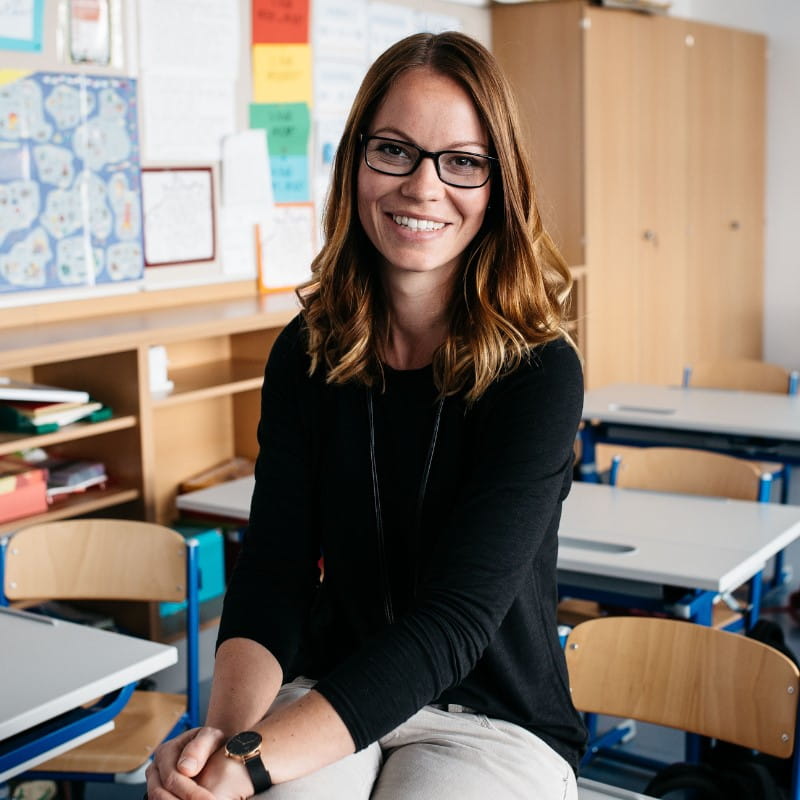 A professional educator seated in her classroom, confidently posing for a photo as she showcases her passion for teaching and creating a positive learning environment.