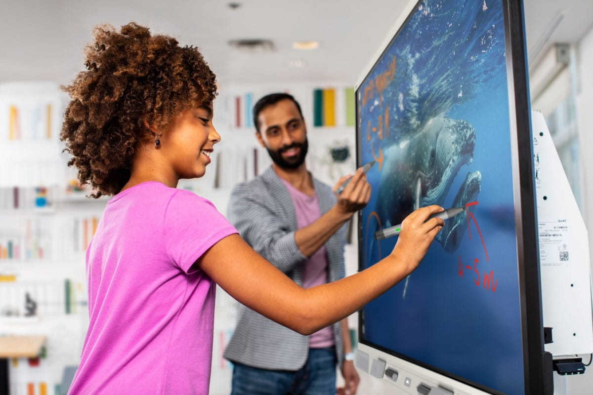 Young girl using the SMART board during an interactive classroom session, with a teacher and classmates engaged in the background.
