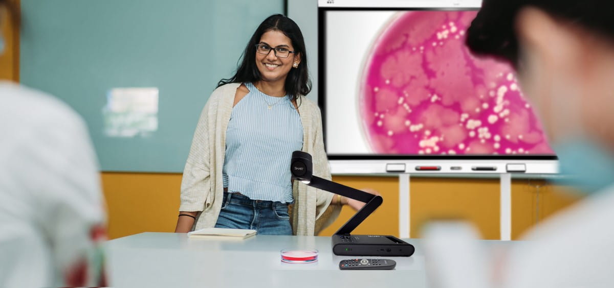 A teacher smiles confidently in a classroom, standing next to a SMART Document Camera projecting a detailed image of cells onto a SMART Interactive Display, engaging students in a biology lesson.