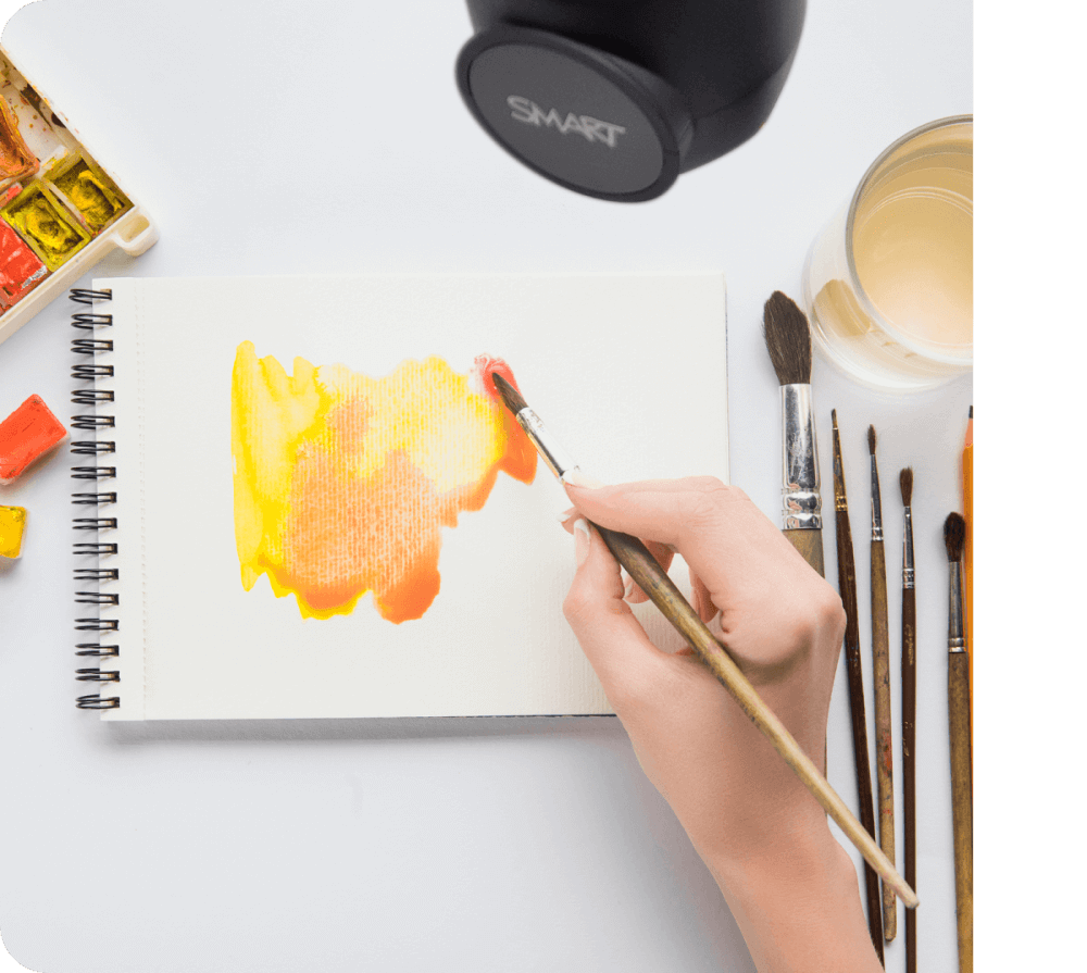 An artist's hand painting a vibrant orange and yellow watercolor on a sketchpad, with a SMART Document Camera positioned above to capture the artwork in real-time for digital sharing or display.
