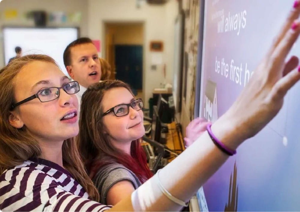 Two students and a teacher are engaging with a SMART Board M700 interactive whiteboard in a classroom setting, demonstrating its object awareness feature with one student touching the screen and the teacher observing attentively.