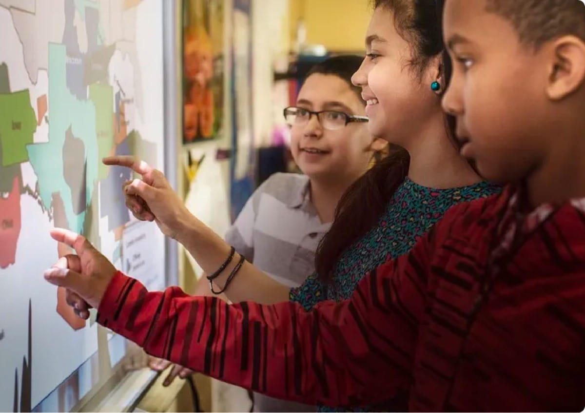 Three enthusiastic students interacting with a SMART Board interactive whiteboard in a classroom, demonstrating dual-user collaboration with simultaneous tool differentiation as they engage with a colorful map.