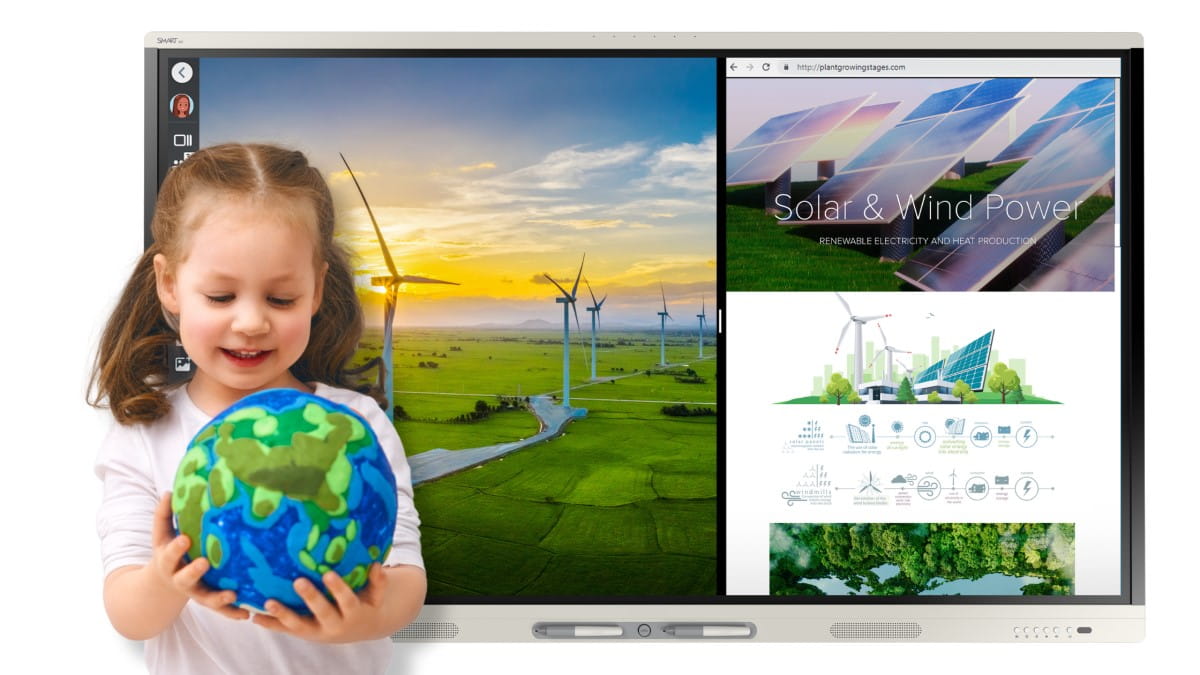 A young girl smiling while holding a globe with illustrations of greenery and water, with a SMART Board displays in the background showing renewable energy themes, one with wind turbines at sunset and another with solar and wind power infographics.