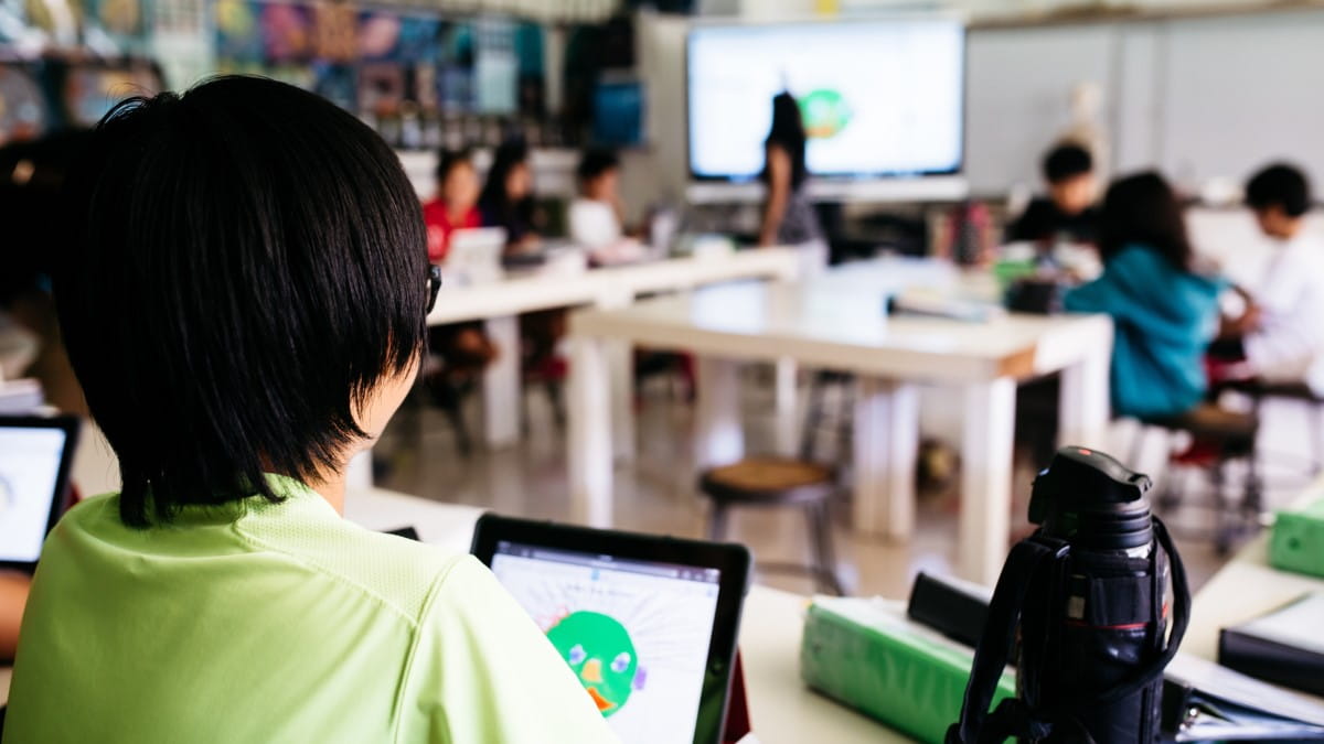 The view from over a students shoulder, showing the Lumio activity on his tablet, as well as a bustling classroom in front of him.