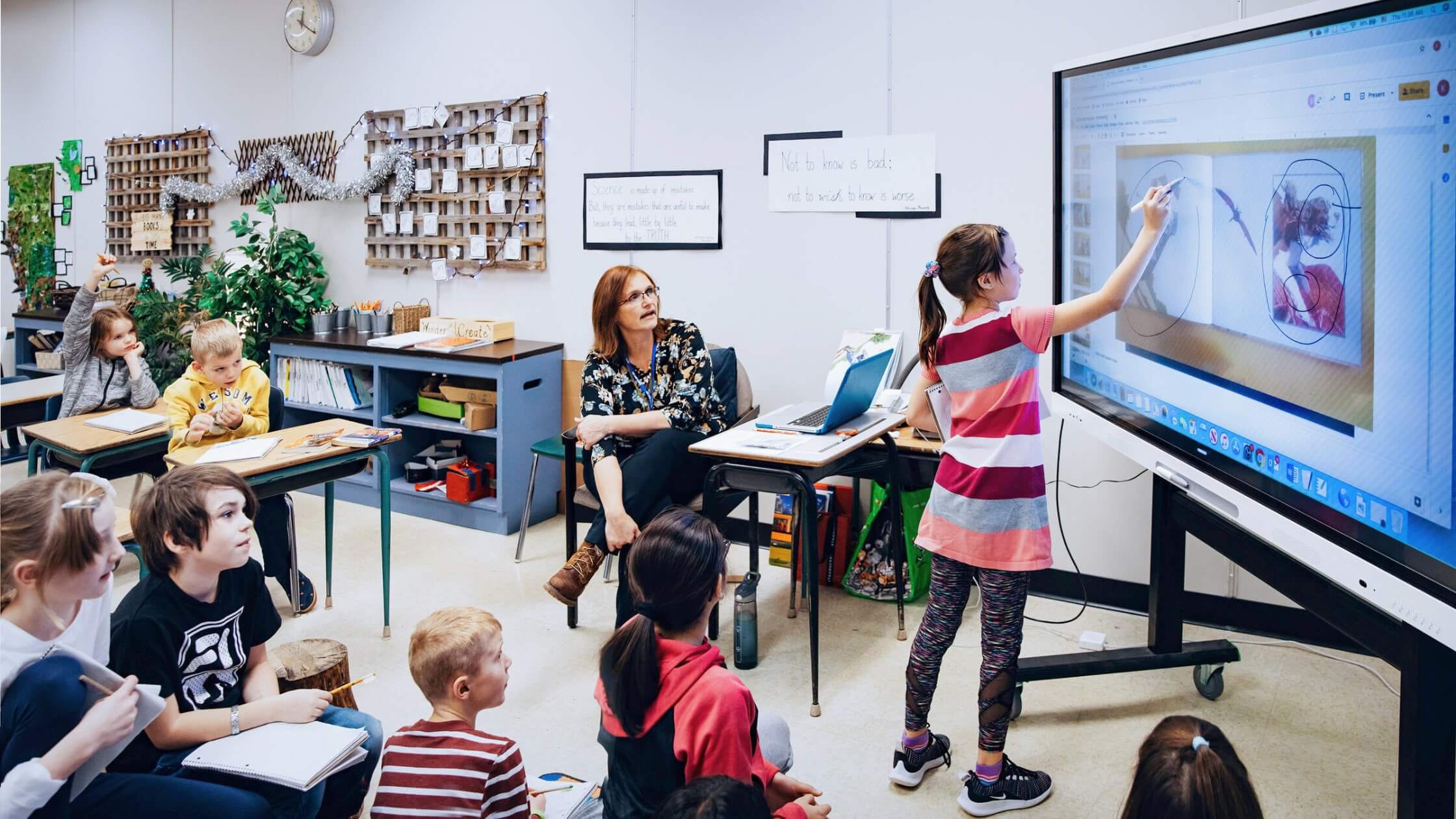 A student engaging with content on a SMART interactive display, while her teacher and classmates watch and support.