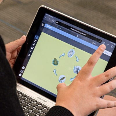 A touch screen laptop showing a Lumio lesson on the screen, while the hands of a student interacts with the content in the lesson.