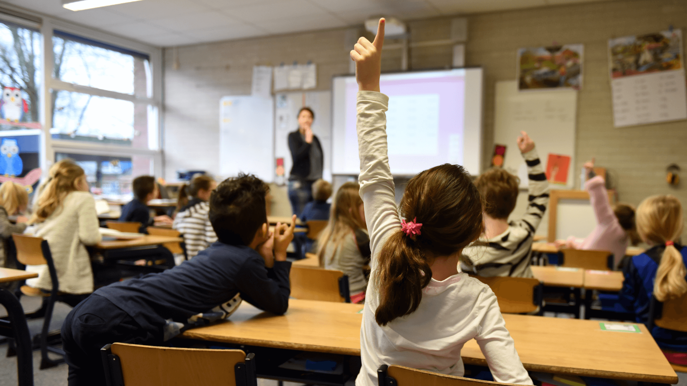 A full classroom participating in discussion with one student raising her hand to answer her teachers question.