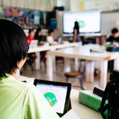 The view from over a students shoulder, showing the Lumio activity on his tablet, as well as a bustling classroom in front of him.