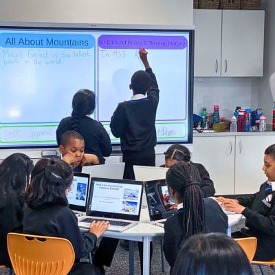 Two students working at a SMART interactive display, while the rest of their group works independently on their student devices at a table in front. 