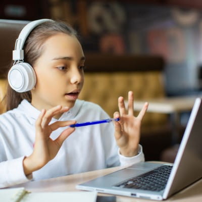 Young girl with headphones using a laptop while holding a pen in hand, having an online learning session or virtual classroom.