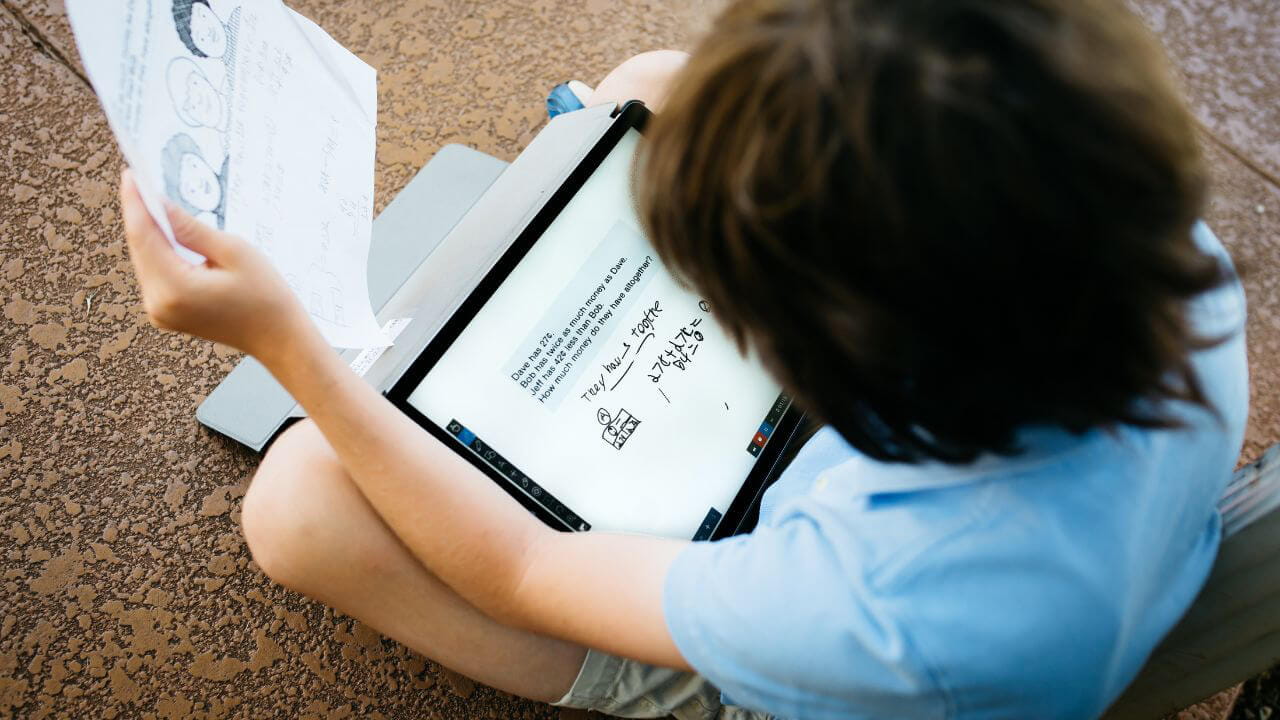 A school aged boy sits cross-legged, working independently on a tablet with paper materials to support.