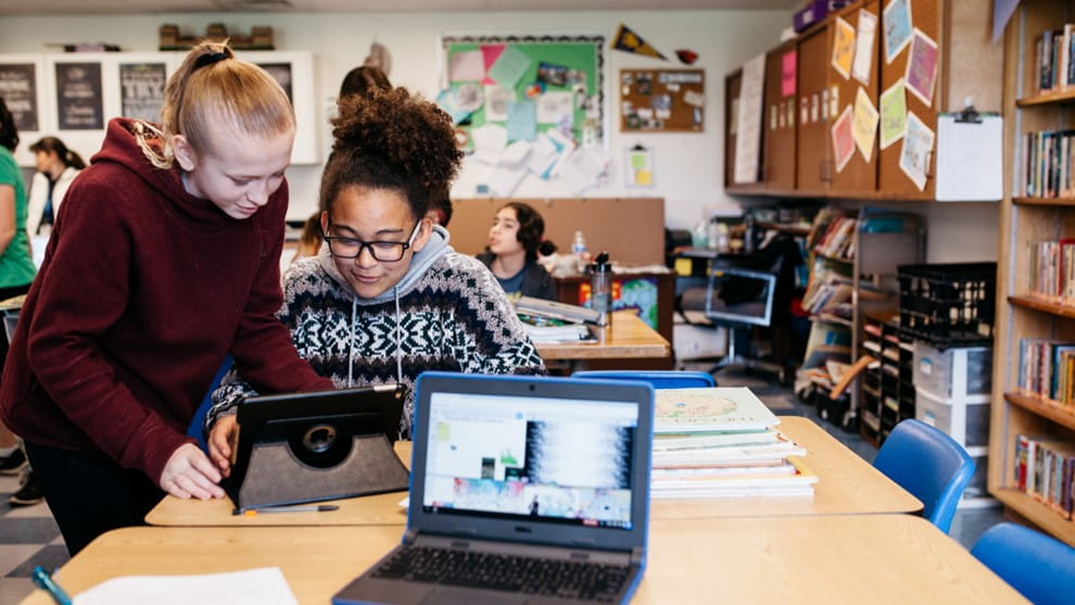 The image depicts two students working together on a tablet device. They appear to be engaged in a collaborative activity, possibly working on a project or studying together