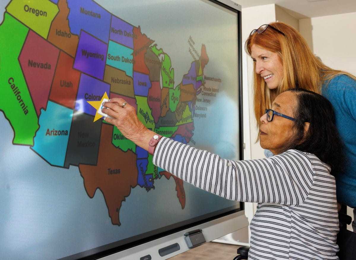Senior care resident and facilitator engaging with a geography lesson on a SMART board to encourage movement.