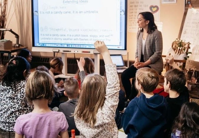 A classroom scene with young students eagerly raising their hands to answer a question, with a teacher next to a SMART board displaying a lesson on extending ideas.