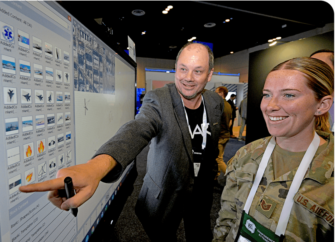 Two individuals, one in military attire, engaging with a SMART Board during a demonstration at an exhibition, showcasing the board's collaborative features.