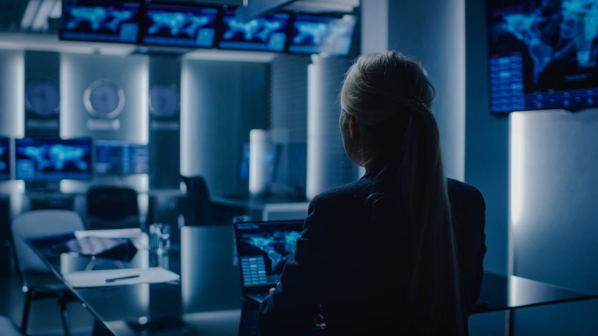 Back view of a woman in a modern control room filled with multiple displays showcasing global networks, emphasizing secure collaboration and decision-making for government agencies.