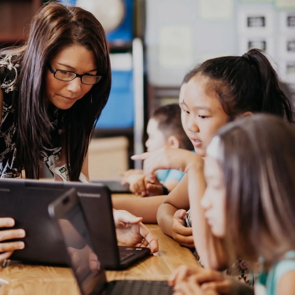 A teacher with glasses leans over to assist a group of young students who are engaging with educational content on a tablet. The children, appearing focused and curious, are pointing at the screen as the teacher guides them through the lesson.