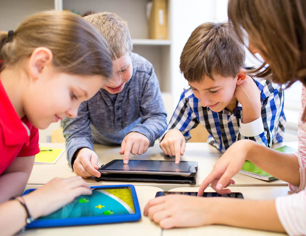 A group of four children in a classroom focused on learning activities using tablets. Two students are sharing a tablet on the right, while another student uses a tablet individually on the left.
