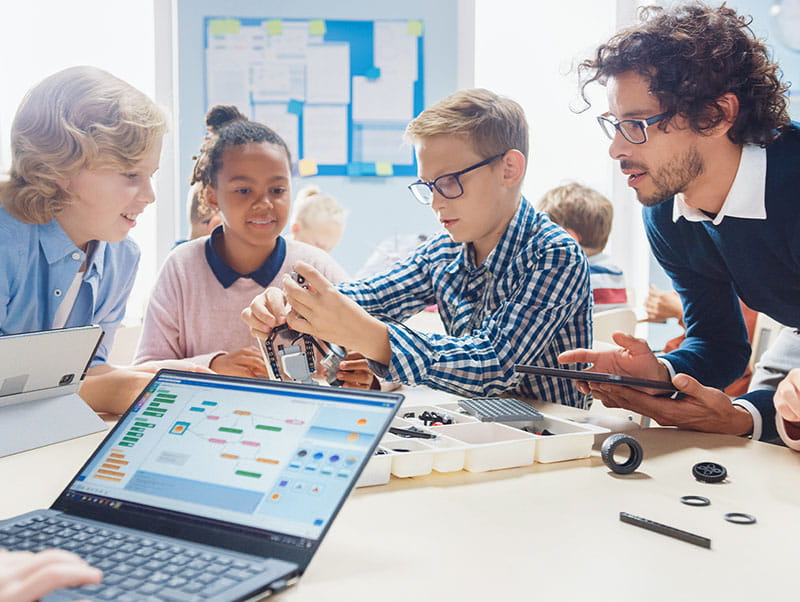 Students and a teacher engaged around a table with a laptop, showing active participation and collaborative learning.