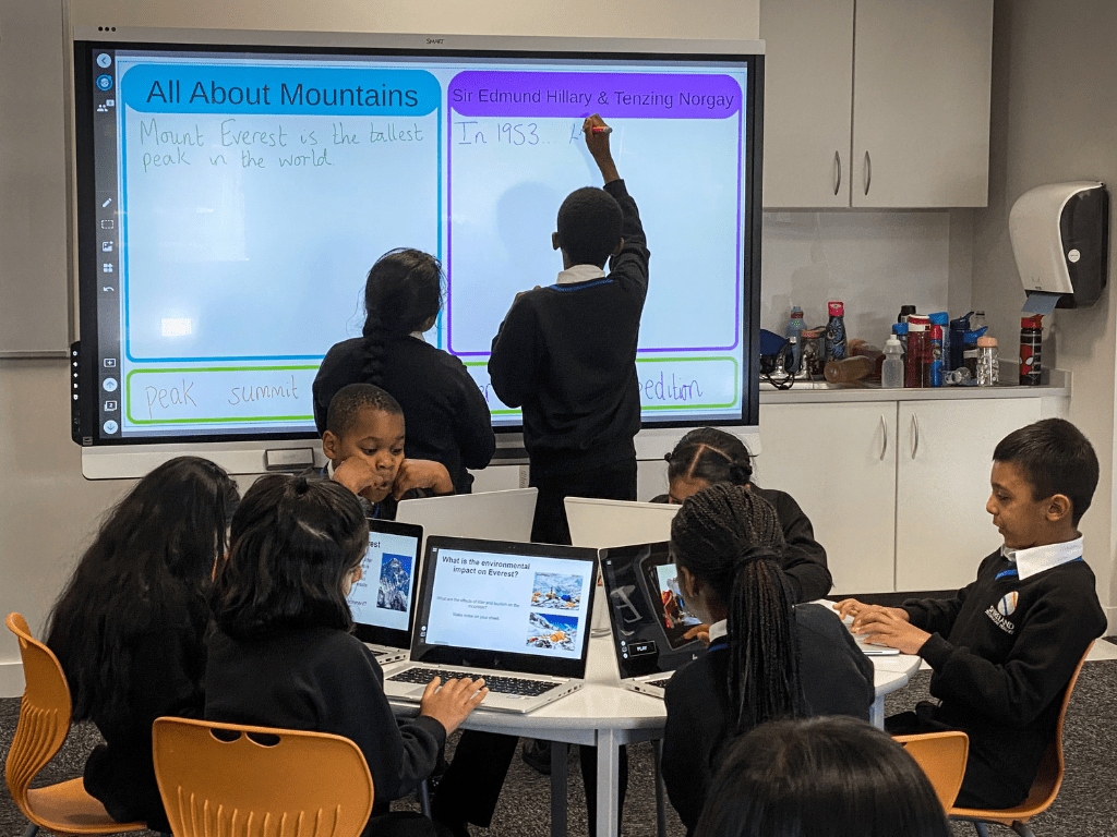 A group of 8 students working on student devices and an interactive display