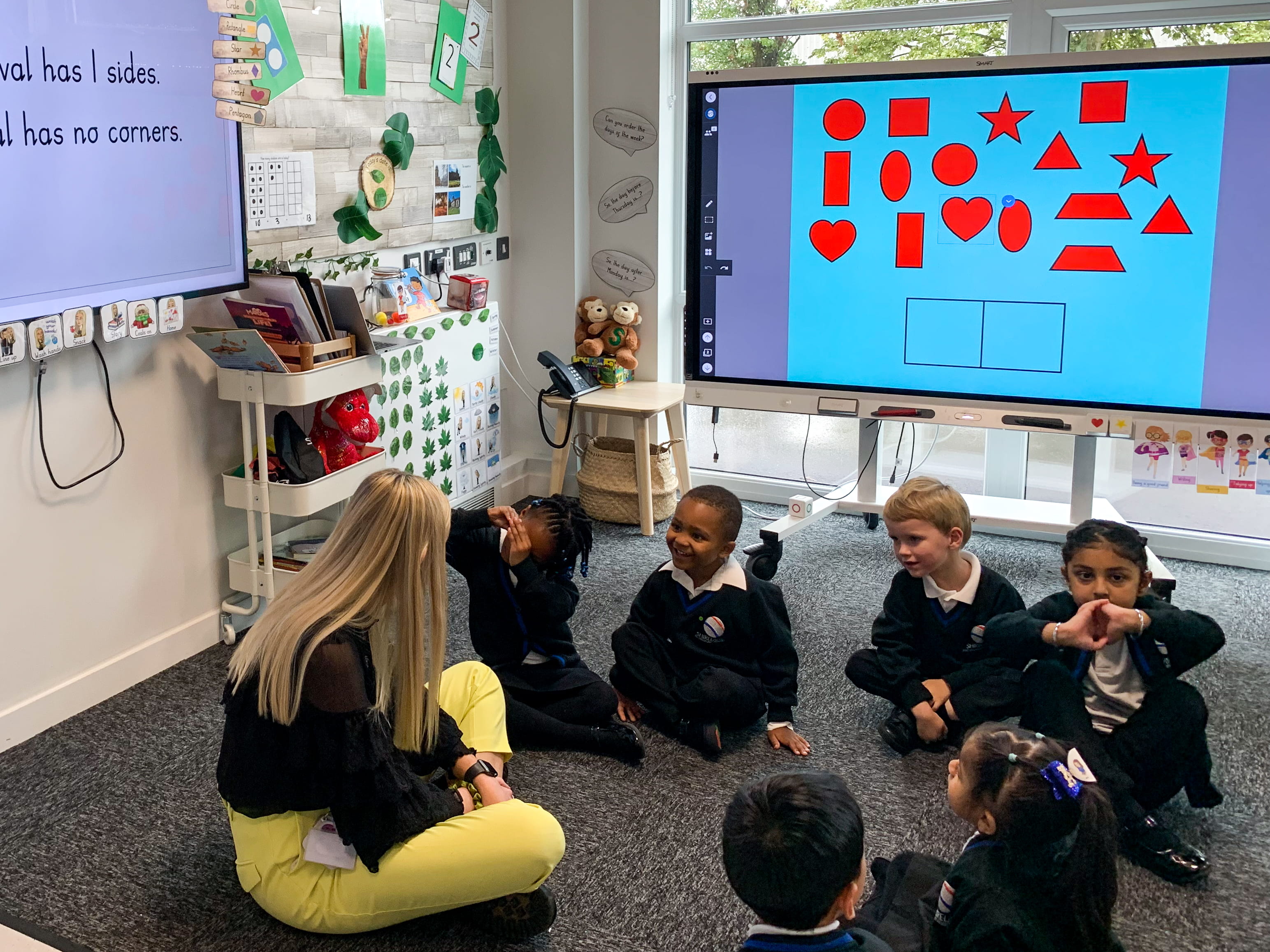 Students gather on the floor with their teacher for a lesson. SMART displays pictured in the background.