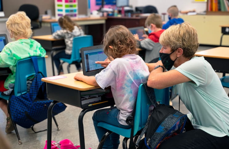 Teacher assisting a young student with a tablet in a classroom.