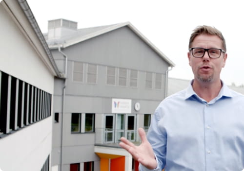 Educator explaining in front of a gray school structure with large windows.