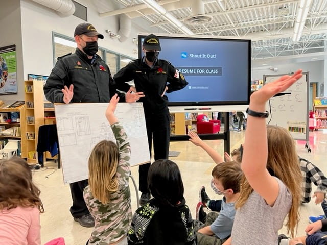  Two officers interacting with young students in a classroom with a SMART Board displaying 'Shout It Out!'.