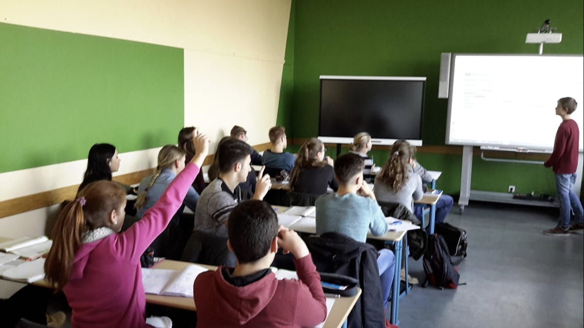 A classroom scene with students seated at desks, looking attentively towards a SMART board where a teacher is presenting.