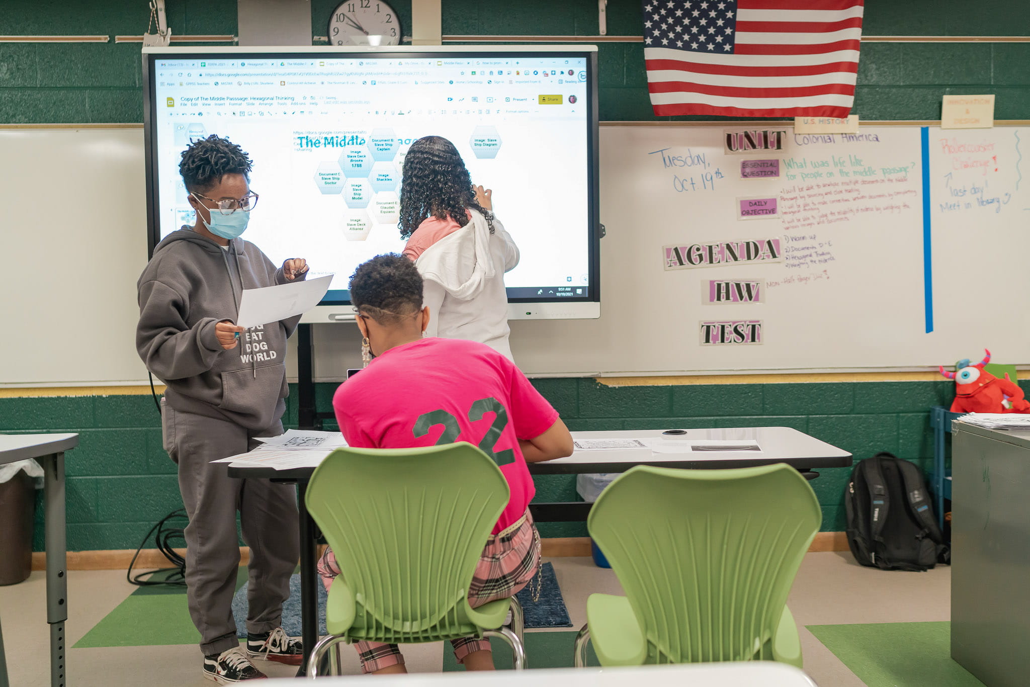 Three students work at a SMART Board in a classroom, with two standing and interacting with the display, while one sits at a desk in a bright pink shirt, focusing on their work.