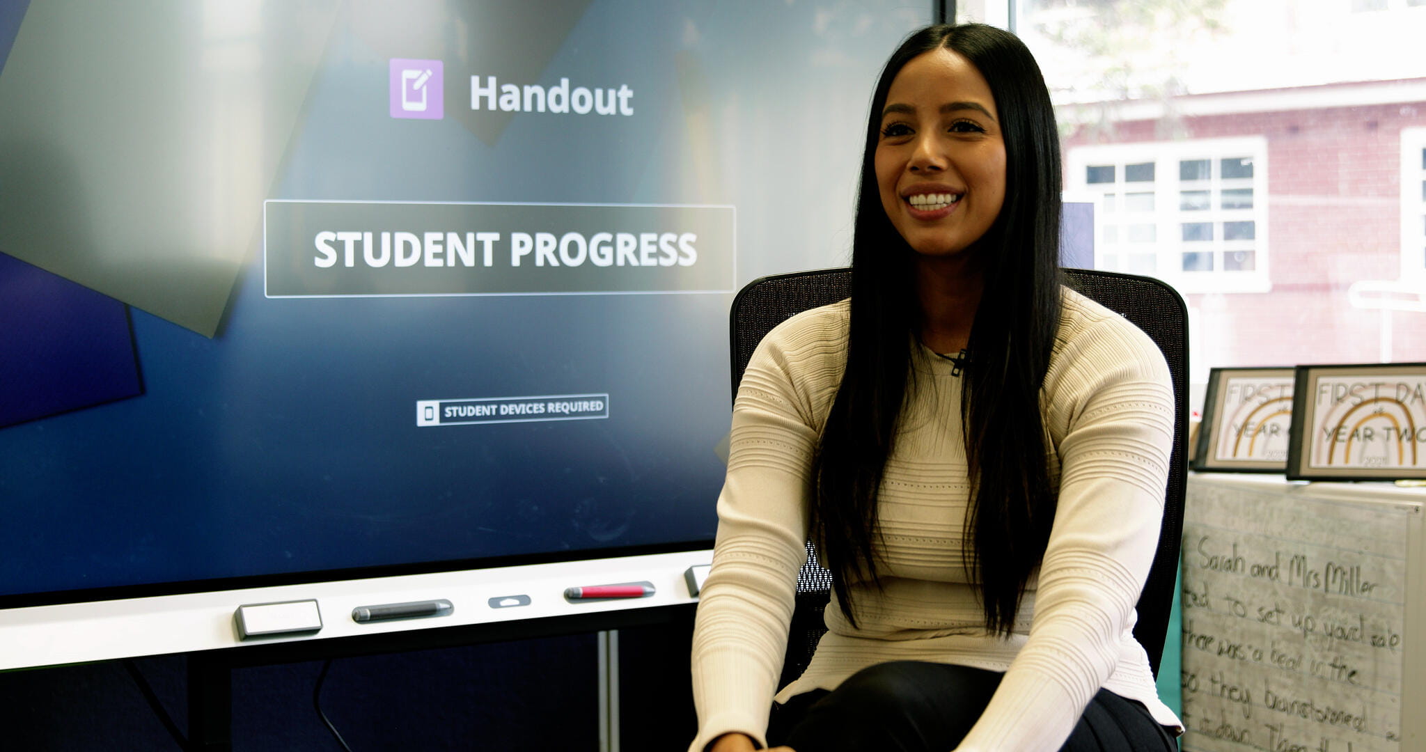 A smiling woman sits in front of a SMART Board displaying 'Handout: Student Progress' on the screen.