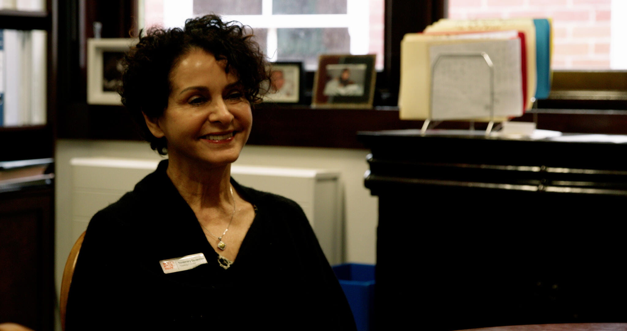 A school administrator smiles while seated in an office with framed photos and certificates in the background.