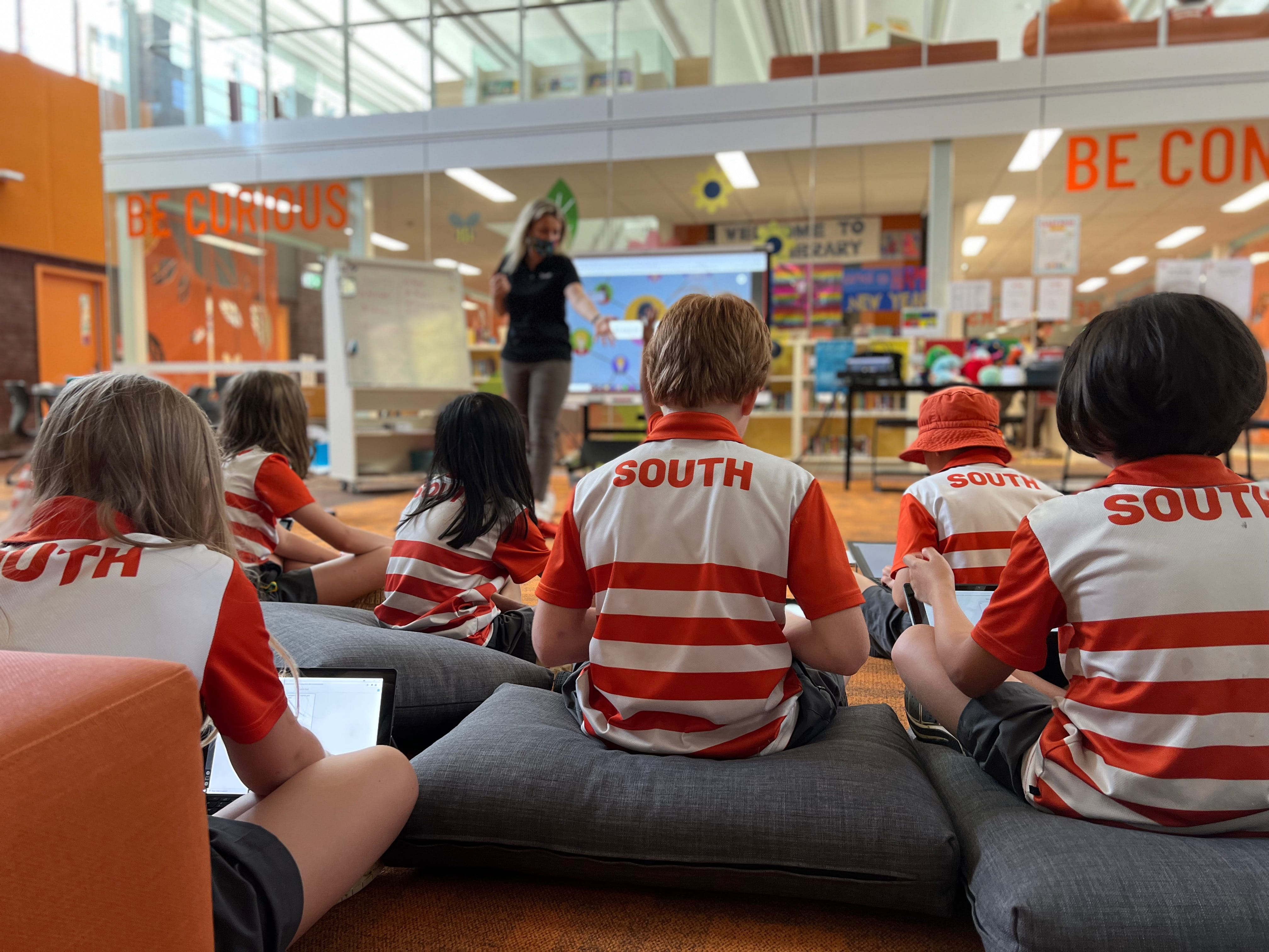 Students in red and white 'South' uniforms sit on cushions in a library while a teacher conducts a lesson on a SMART Board.