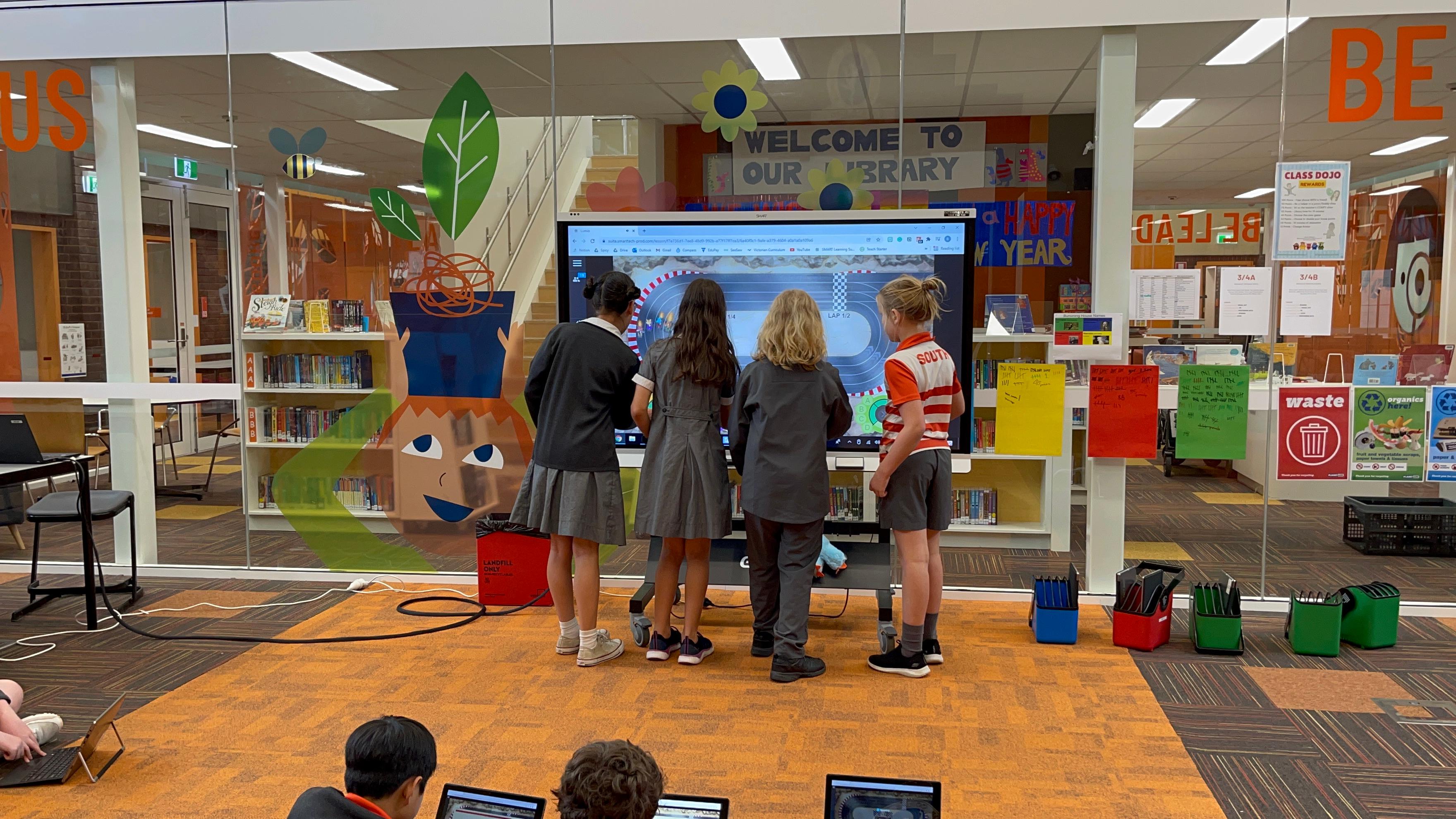 Four students gather around a SMART Board in a brightly decorated library, exploring an interactive activity.