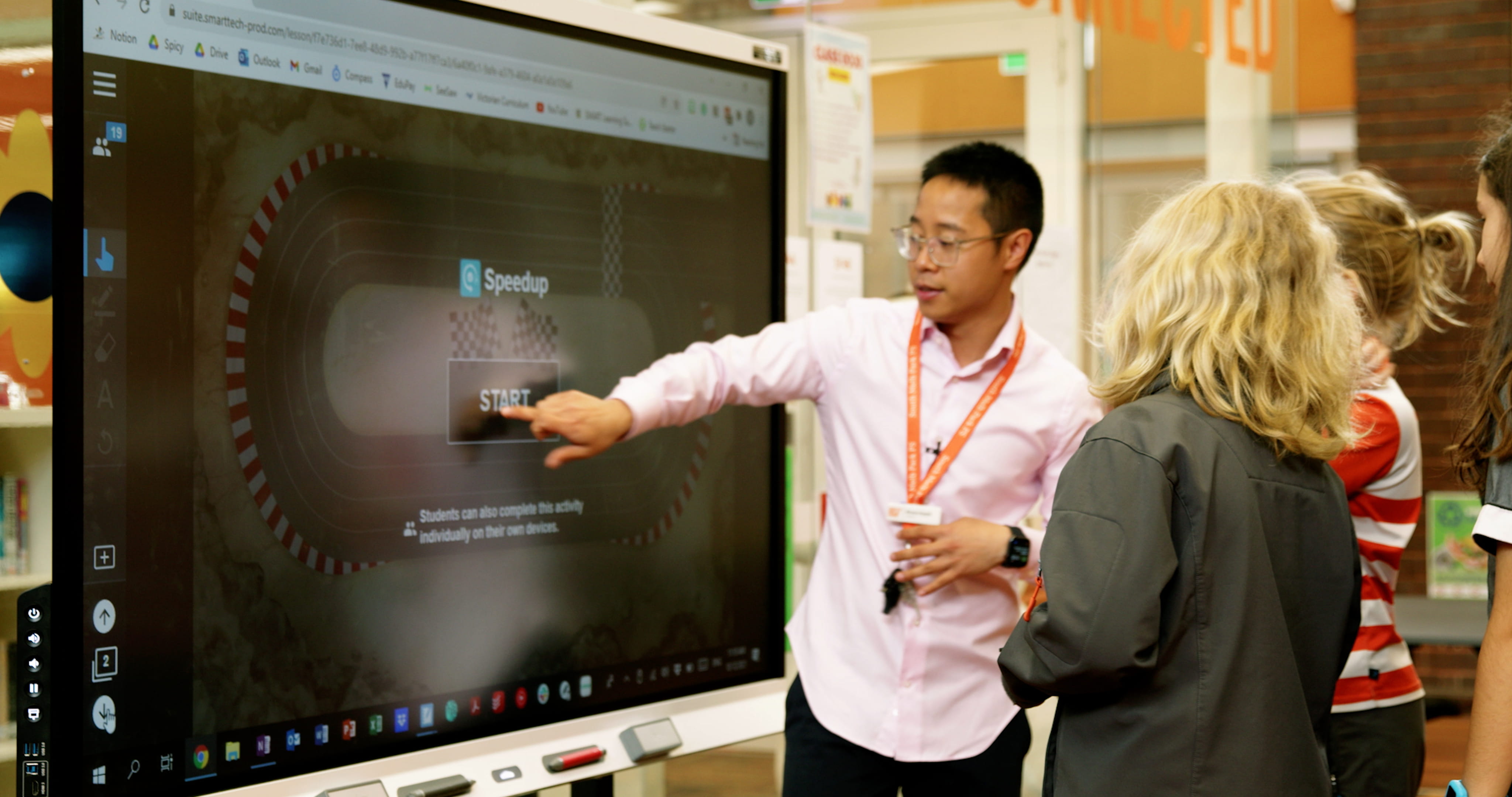 A teacher in a pink shirt points to an interactive activity on a SMART Board while students watch attentively.
