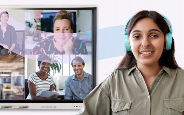 A woman in a casual outfit smiles engagingly in front of a SMART board showing a video call with colleagues, symbolizing remote teamwork and communication.