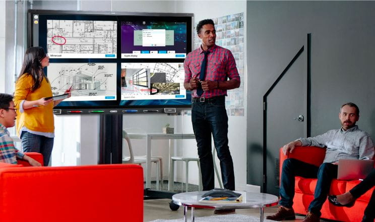 A modern office meeting where a professional stands by a SMART Interactive display, presenting to seated colleagues, showcasing the integration of technology for efficient workflow.