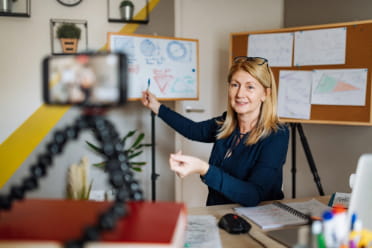 Professional woman recording a video presentation using a smartphone on a tripod, explaining a flowchart in an office setting.