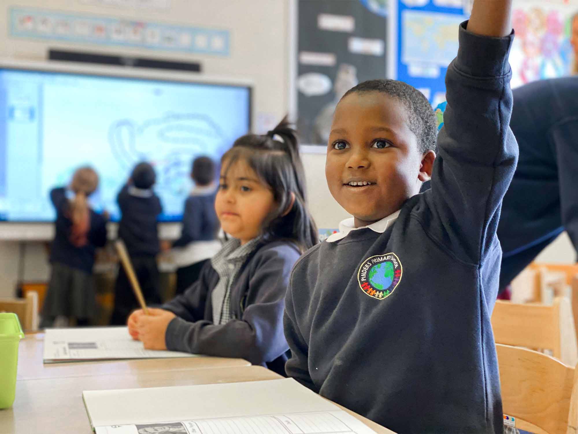 A young boy in a school uniform excitedly raises his hand in a classroom with other students, interacting with a SMART board in the background, demonstrating active learning and engagement with technology.