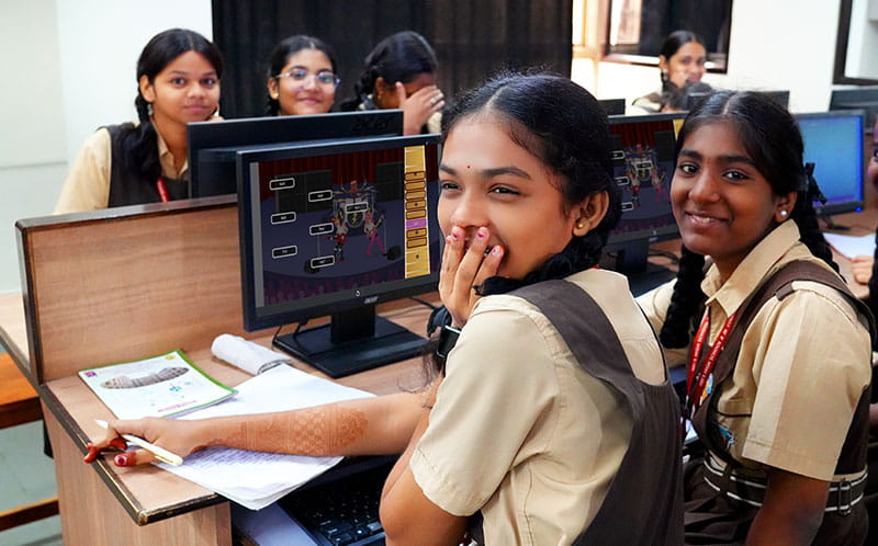 Students in a computer lab smiling and engaging with each other during a fun and interactive session.